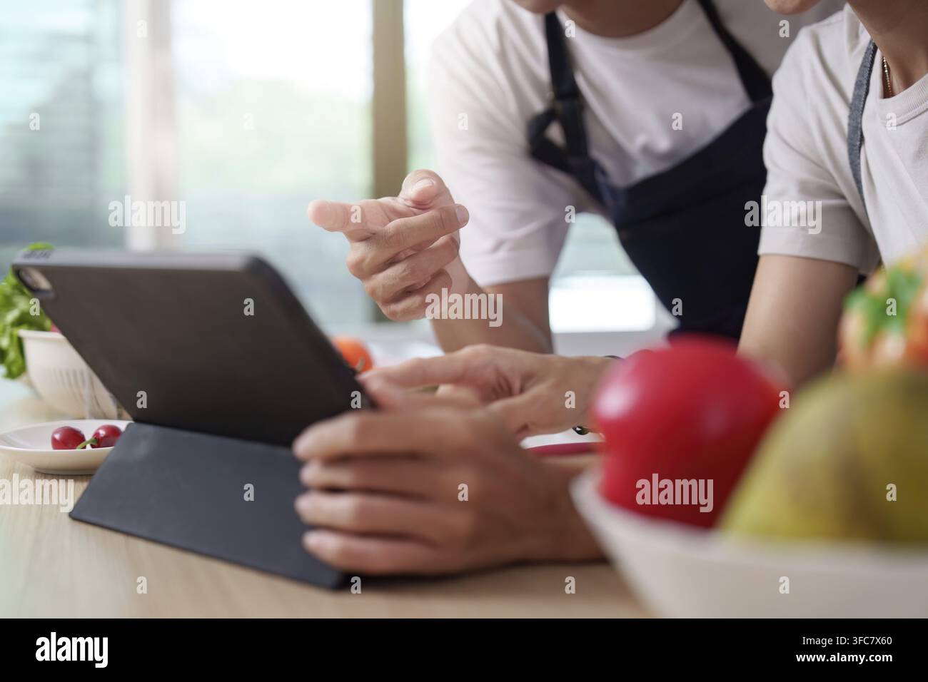 Ein Paar arbeitet mit einem Tablet und recherchiert neue Rezepte, während es in einer modernen Küche kocht. Der Fokus beim Kochen liegt auf Technologie Stockfoto