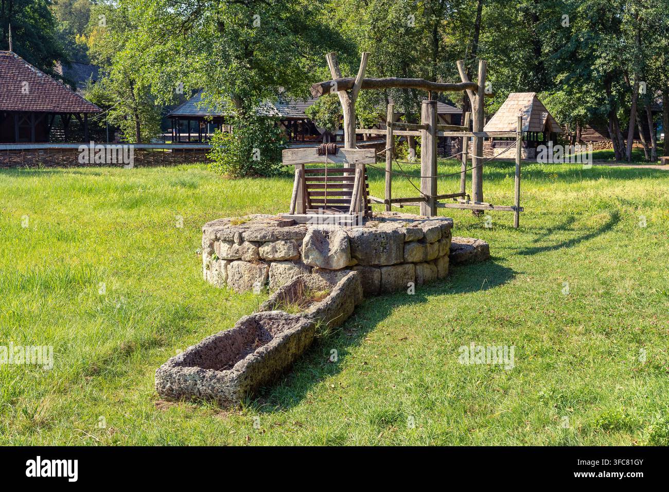 Alter steinerner Wasserbrunnen mit hölzerner Winde und Tränke in einer grasbewachsenen ländlichen Umgebung Stockfoto