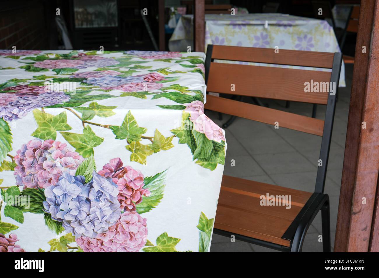Gemütlicher Außentisch eines Cafés, bedeckt mit einer weißen Tischdecke mit einem hellrosa und lila Hortensienblumenmuster. Nahaufnahme eines rustikalen Holzstuhls Stockfoto