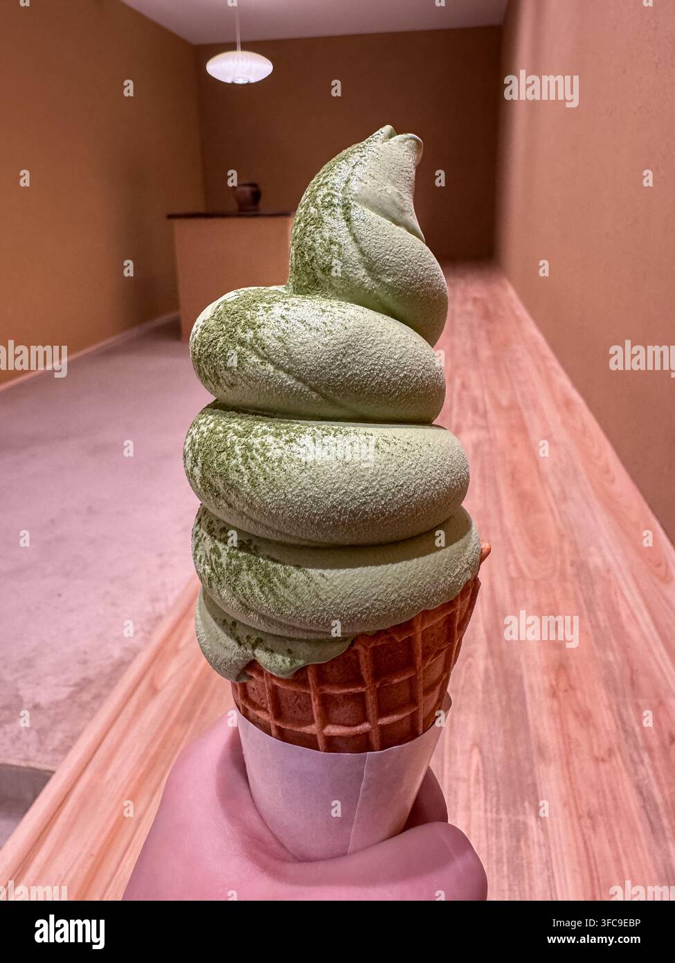 Matcha-Grüntee-Softeis in einer Waffeltasche, die von einer Hand gehalten wird, vor einem minimalistischen rosa Interieur. - Smartphone-aufgenommenes Stockfoto