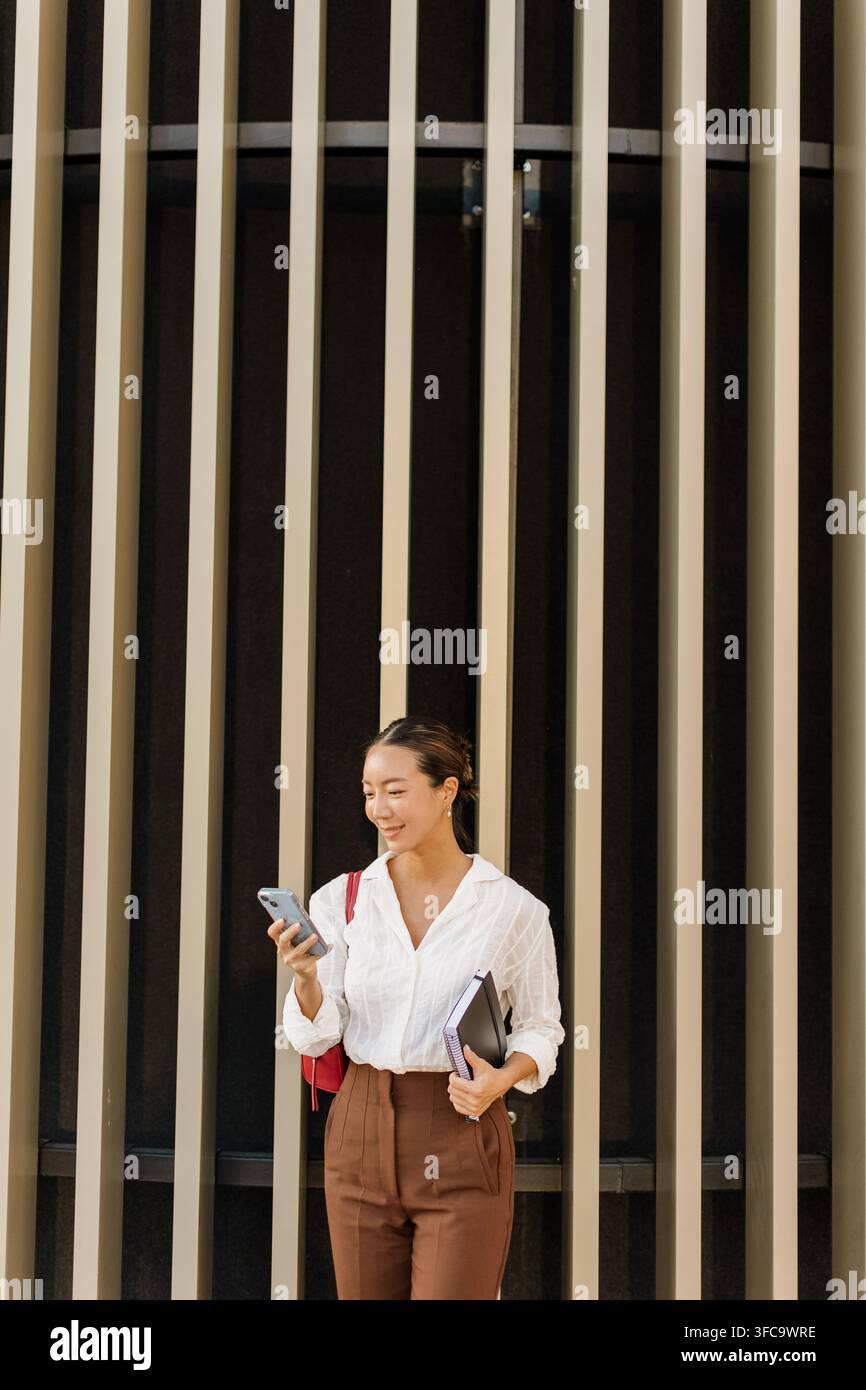 Eine fröhliche Koreanerin steht vor der gestreiften Wand eines modernen Gebäudes, lächelt, während sie ihr Handy überprüft und ein Notizbuch hält. Stockfoto