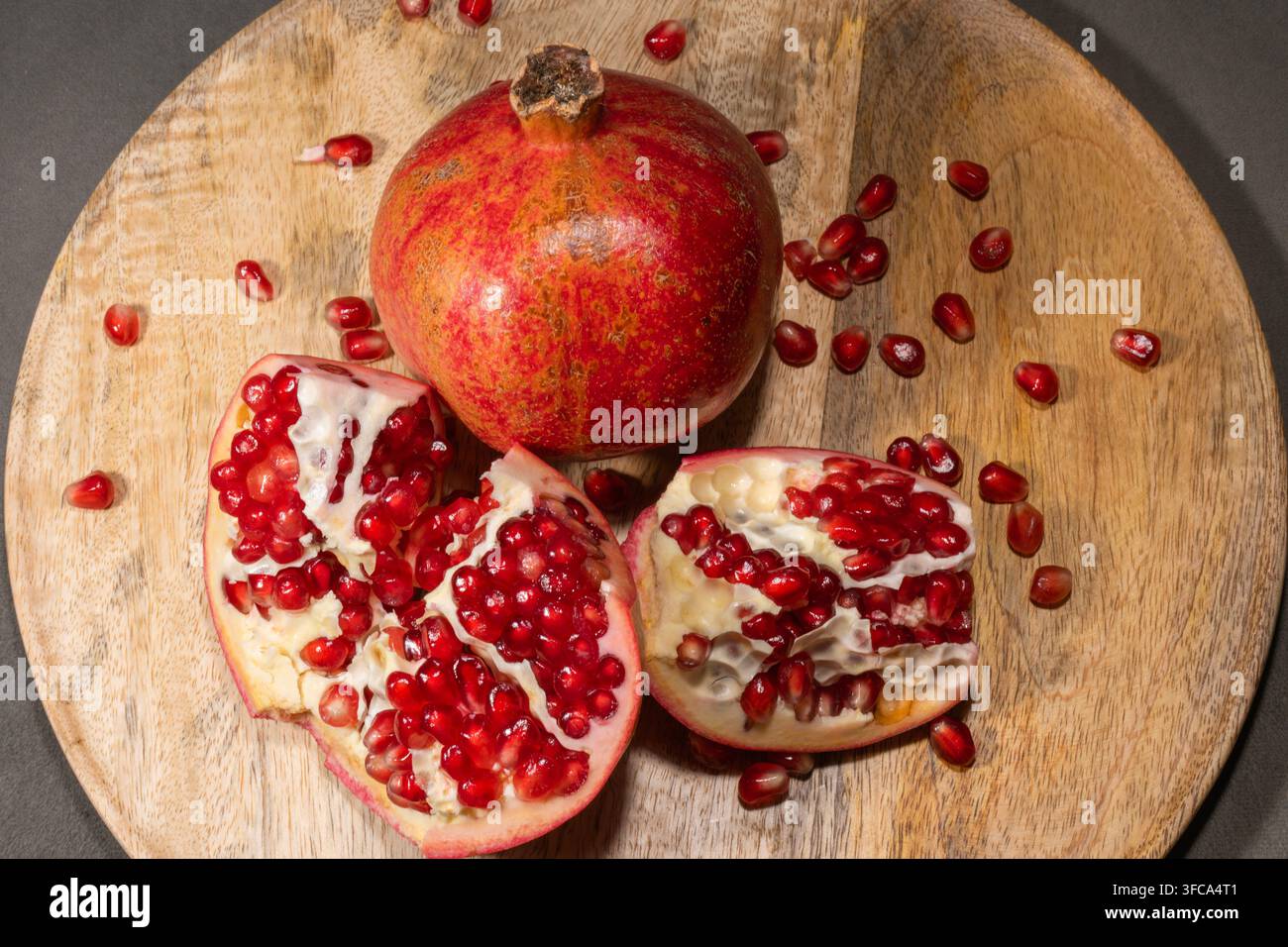 Ein ganzer Granatapfel und mehrere geschnittene Teile zeigen lebhafte rote Arillen, die über eine rustikale Holzschale verstreut sind. Stockfoto
