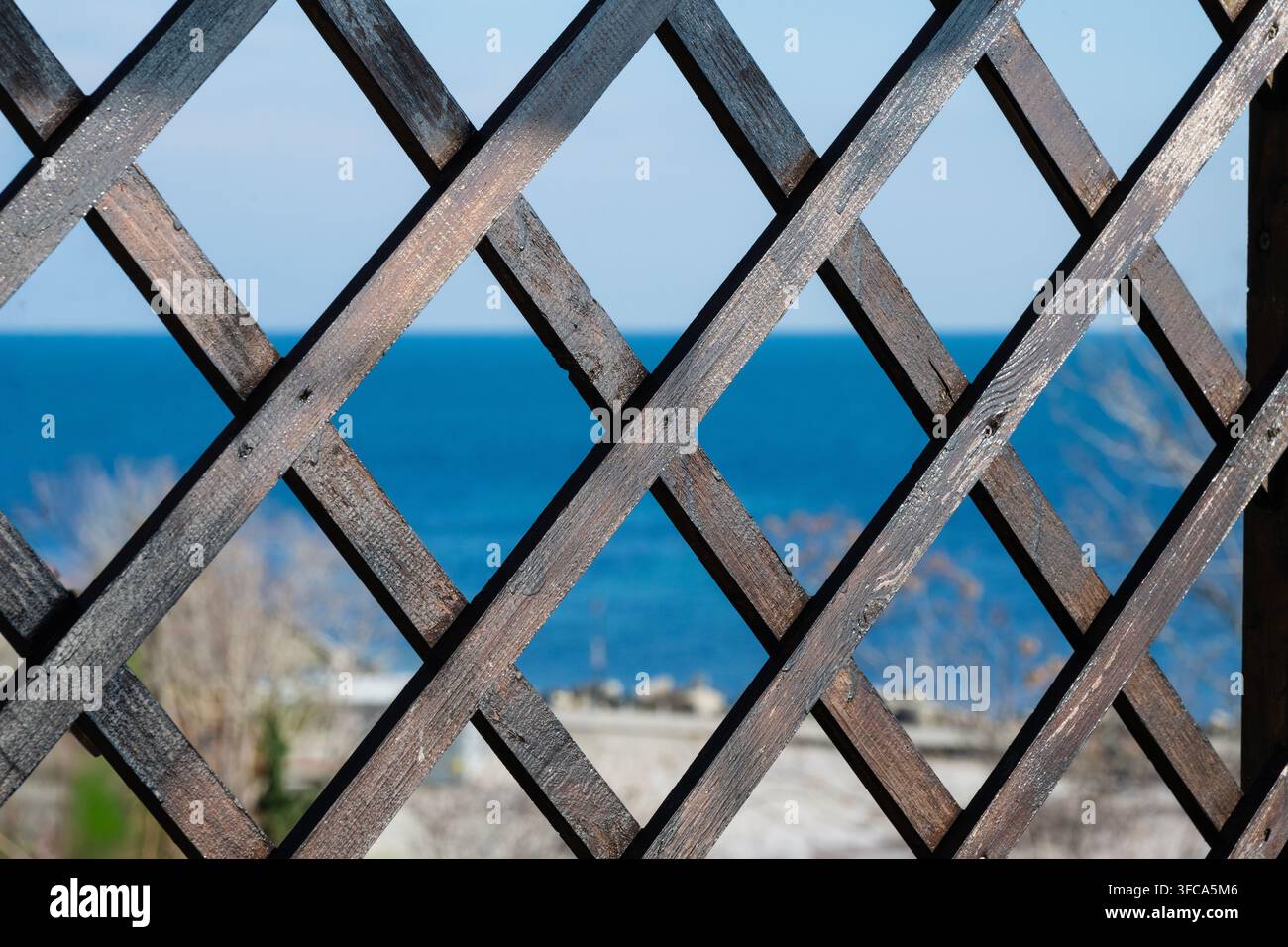Ein dunkler hölzerner Kreuzgitterzaun umrahmt eine verschwommene malerische Aussicht auf das lebendige blaue Meer und den klaren Himmel an einem hellen, sonnigen Tag. Stockfoto
