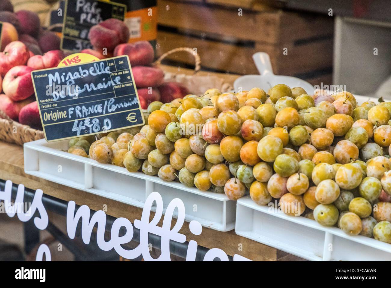 Frische Mirabellen liegen zum Verkauf an einem Marktstand in der Rue Braconnot in Nancy, Grand Est, Frankreich, mit einem Schild, das die geschützte Ursprungsbezeichnung „Mirabelle de Lorraine“ nennt. Die kleine gelbe Pflaume hat seit 1995 den Status einer geschützten geografischen Angabe (g.g.A.) der EU; die Lorraine produziert etwa 90 Prozent des weltweiten Mirabellenertrags. Im Hintergrund sind Pfirsiche gestapelt. Stockfoto
