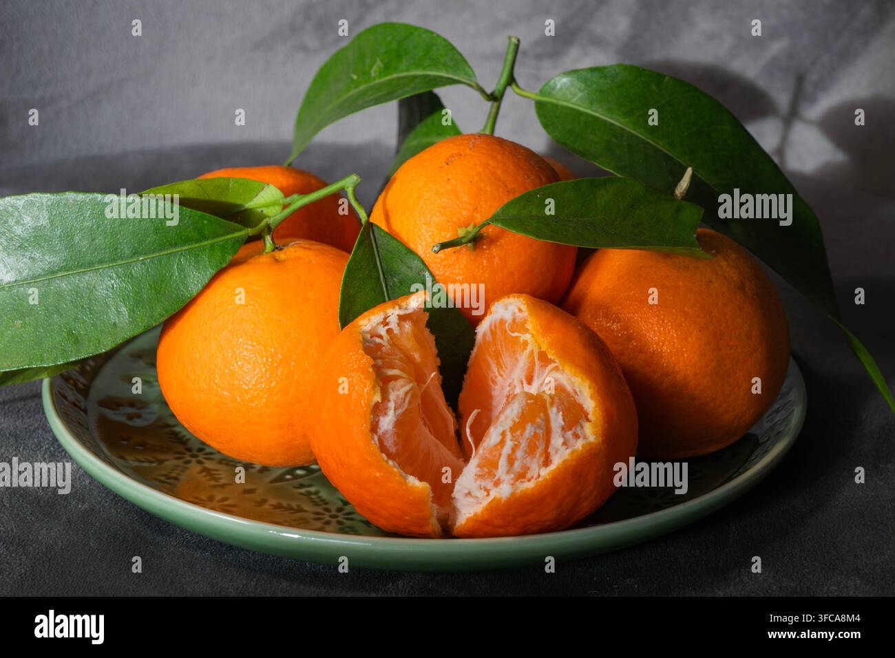 Frische reife Mandarinen mit grünen Blättern liegen auf einem dekorativen Teller, darunter eine aufgeschnittene, die ihre Segmente offenbart. Stockfoto