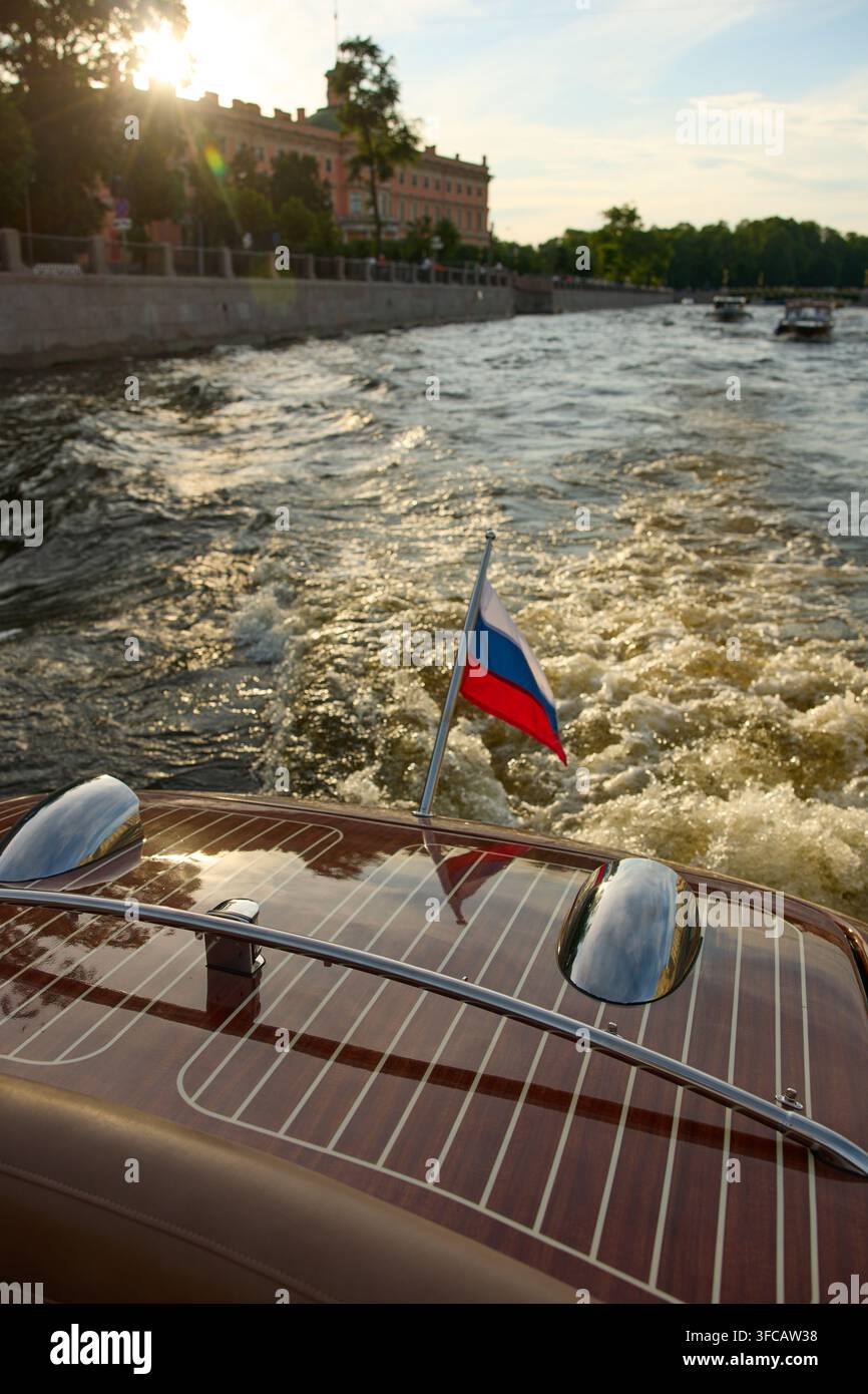 der glänzende Heck eines Vintage-Vergnügungsboots bei Sonnenuntergang, das Boot in Bewegung, blubberndes Wasser unter dem Boot, die Flagge Russlands weht im Stockfoto