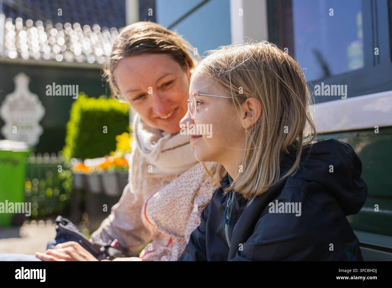 Eine Mutter und ihre Tochter sitzen zusammen auf einer Bank, während sie die Niederlande erkunden. Sie teilen ein Lächeln in einer lebhaften Reiseumgebung. Stockfoto