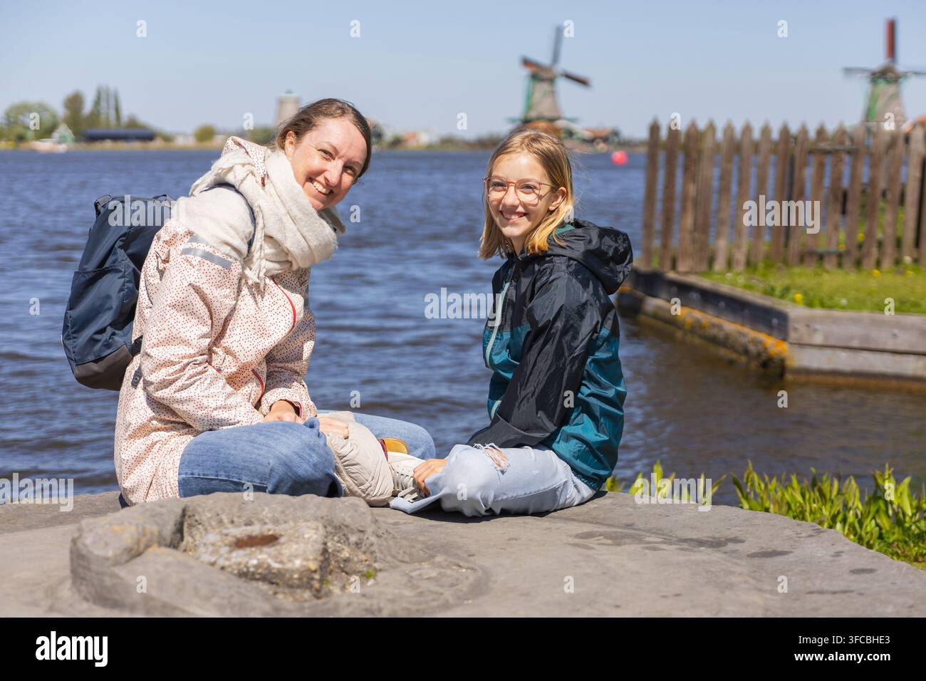 Eine Mutter und ihre Tochter sitzen zusammen am Wasser, genießen ihre Reise in den Niederlanden und blicken auf Windmühlen in der Ferne. Stockfoto