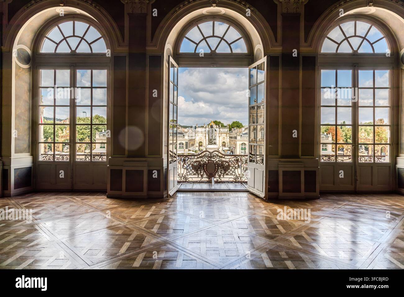 Eine offene Doppeltür in einem mit Holzparkett ausgelegten Salon des Hôtel de Ville de Nancy in Nancy, Grand Est, Frankreich, rahmt den Blick auf den Place Stanislas und die Stanislas-Statue. Ein schmiedeeisernes Balkongeländer rahmt den Ausblick. Stockfoto