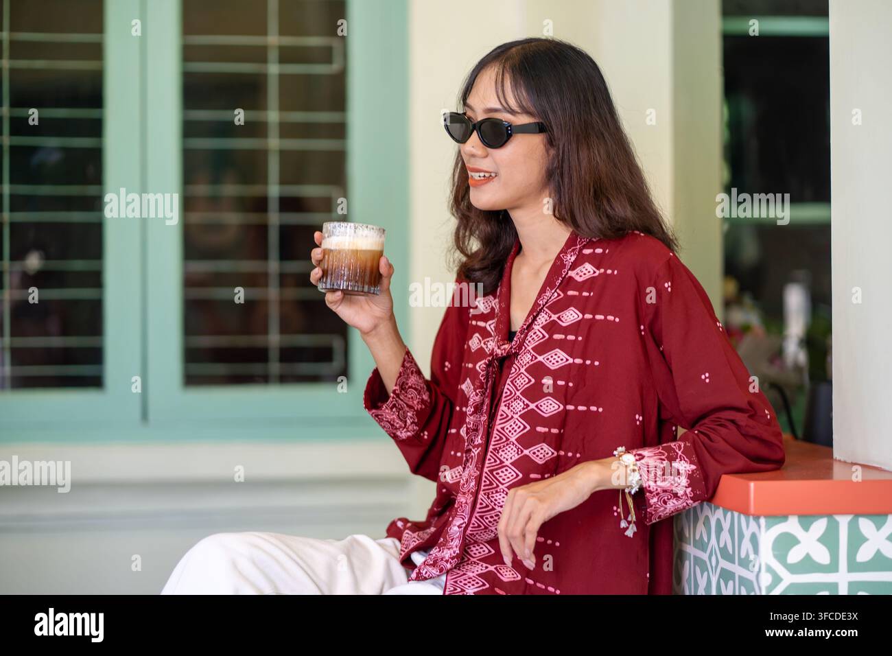 Stylische indonesische Frau trägt eine moderne Batikbluse und eine Sonnenbrille und genießt einen Kaffee, während sie in einem farbenfrohen Café-Interieur entspannt. Stockfoto