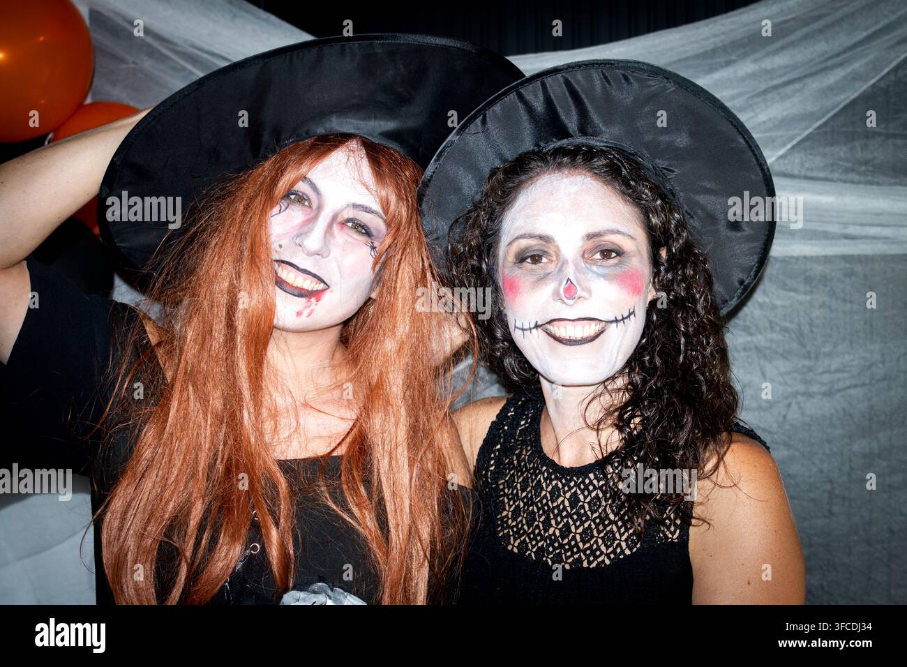 Zwei Frauen, die Hexenhüte und kreatives Halloween-Make-up tragen, lächeln zusammen während einer gruseligen Kostümparty-Feier. Stockfoto