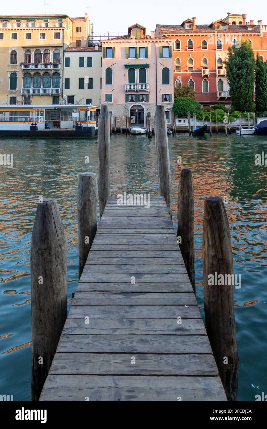 Holzsteg an einem venezianischen Kanal mit farbenfrohen historischen Häusern am Ufer, ruhigem, welligem Wasser und einem kleinen Boot an einem italienischen Stadtabend. Stockfoto