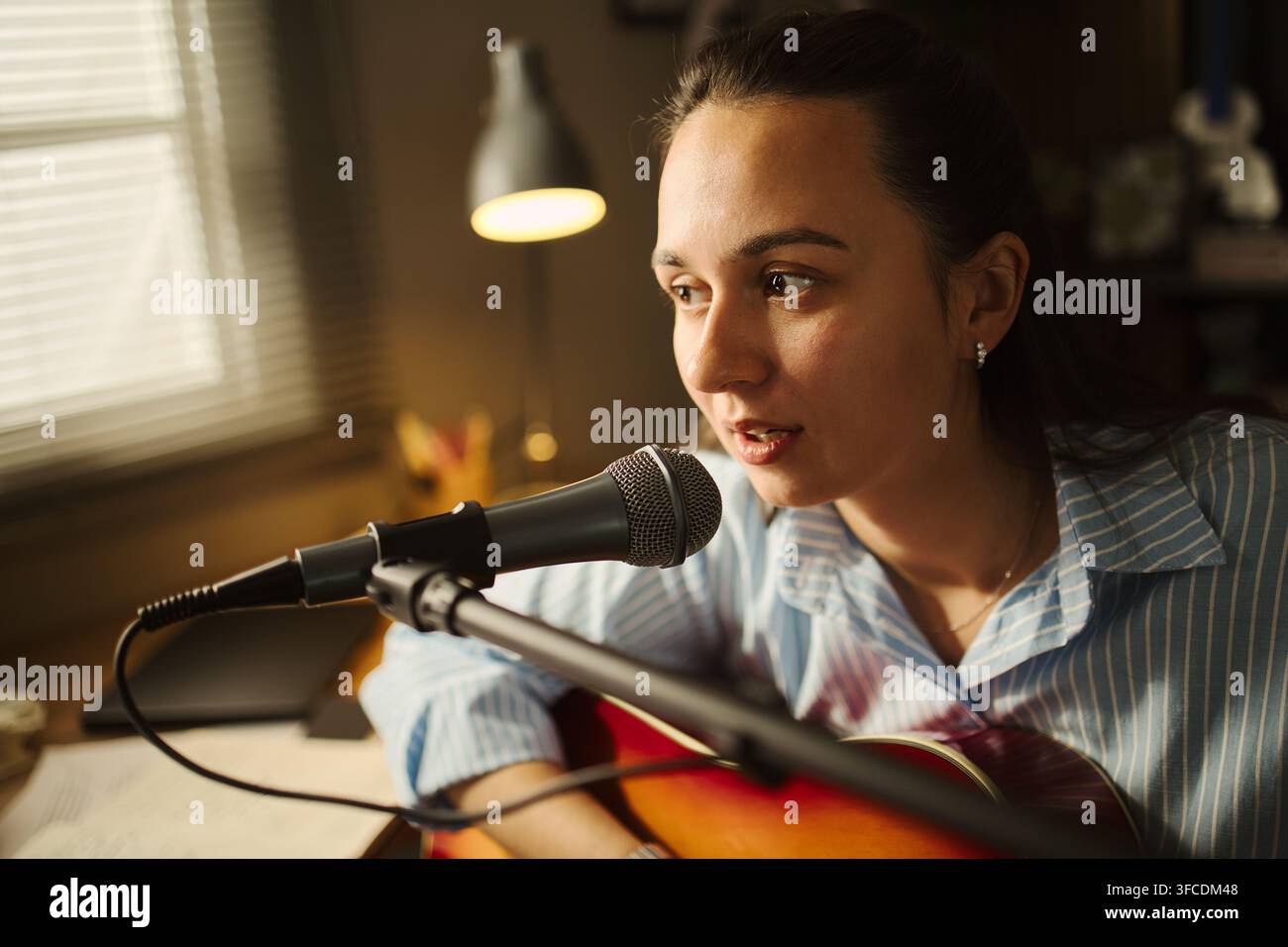 Junge kaukasische Frau singt in ein Mikrofon, während sie in einem Heimstudio Akustikgitarre spielt. Das Bild repräsentiert kreative Inhaltsgestaltung, geeignet für Musikbildungsmarketing Stockfoto