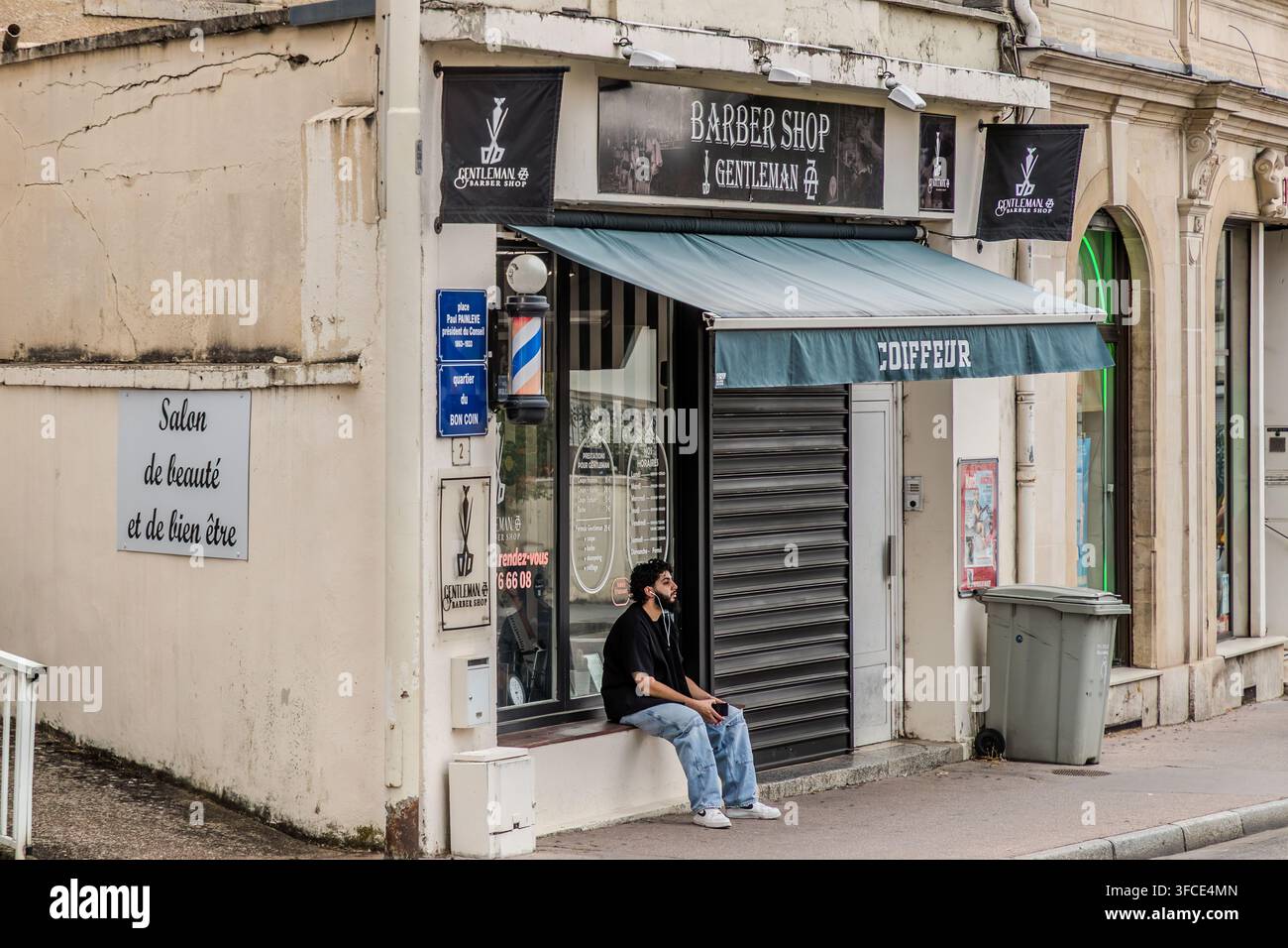 Ein Mann sitzt auf einer Bank vor dem geschlossenen Friseursalon „Barber Shop Gentleman“, der durch einen Friseursturm und ein Vordach mit der Aufschrift „Coiffeur“ gekennzeichnet ist, in Nancy, Grand Est, Frankreich. Ein benachbartes Schild wirbt für einen „Salon de beauté et de bien être“ (Schönheits- und Wellnesssalon). Stockfoto