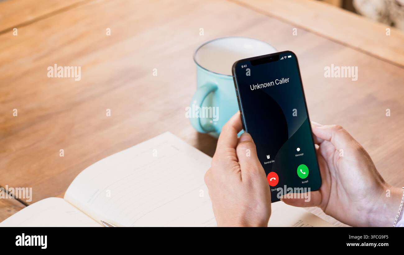 Nahaufnahme von Händen, die ein Smartphone halten, das einen unbekannten eingehenden Anruf anzeigt, neben einem offenen Notizbuch und einer Kaffeetasse auf einem Holztisch. Stockfoto
