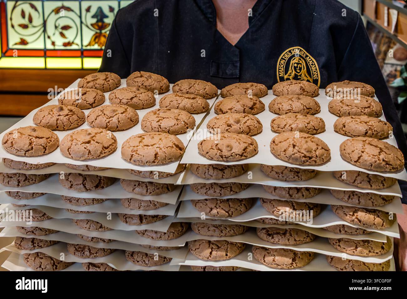 Ein Bäcker aus der Konditorei Maison des Sœurs Macarons präsentiert einen Stapel frisch gebackener Macarons de Nancy auf der Rue Saint-Dizier in Nancy, Grand Est, Frankreich. Die traditionellen Mandelmakronen unterscheiden sich von den bekannteren französischen Macaron-Sandwichkeksen durch ihre gerissene, ungefüllte Kruste. Der Tradition nach suchten Nonnen während der Französischen Revolution Zuflucht im Haus der Konditorei und backten dort die ersten Macarons – historisch nicht zweifelsfrei bestätigt, aber der Ursprung des Namens „Sœurs Macarons“.  Stockfoto