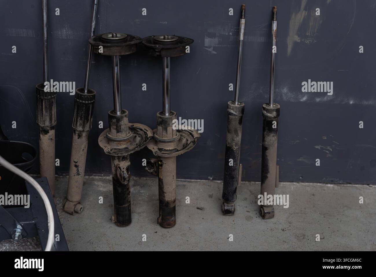 Abgenutzte Federbeinstützen und Stoßdämpfer lehnen an einer gestrichenen Wand, zeigen Rost, Schmutz, beschädigte Stiefel an jeder Einheit. Gruppierte Teile deuten auf eine Werkstatt hin Stockfoto