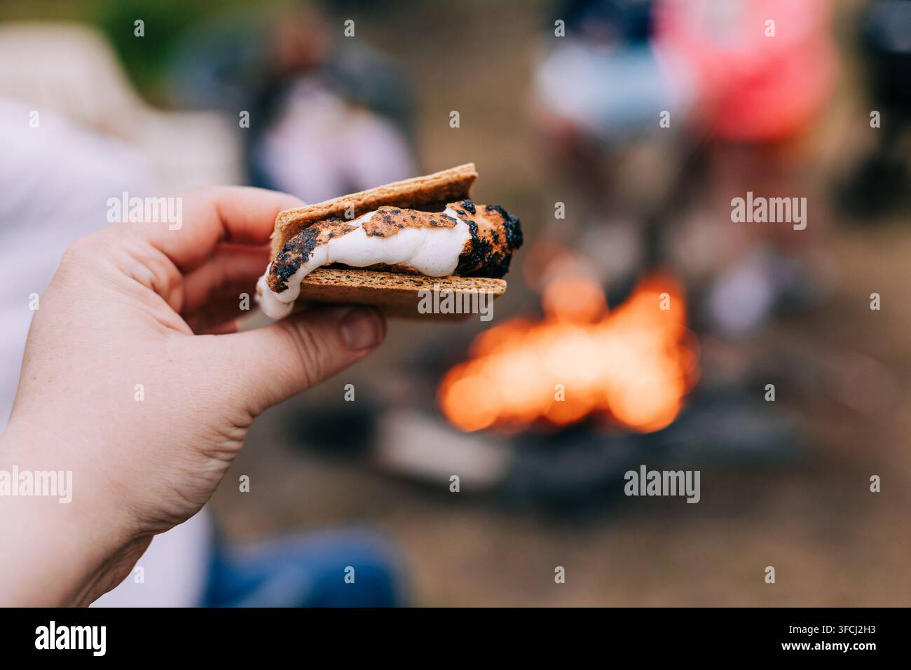 Hand hält ein frisch geröstetes S’more mit geschmolzenem Marshmallow und Graham-Keksen vor einem glühenden Lagerfeuer während eines Campingausflugs im Freien Stockfoto