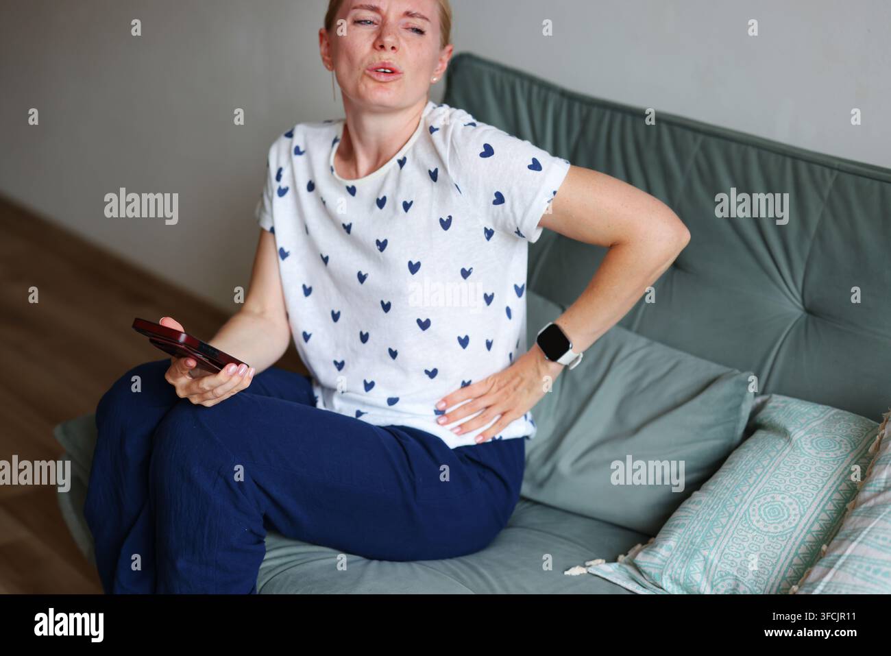 Kranke Frau mit Magenschmerzen, die einen KI-Chatbot auf dem Smartphone für Gesundheitsratschläge nutzt, während sie zu Hause auf dem Sofa ruht. Stockfoto
