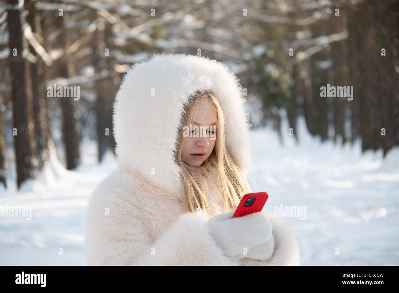Junge blonde Frau in warmem weißem Mantel, die in einem verschneiten Wald ein Smartphone benutzt, Winterkommunikation und mobiler Lebensstil im Freien. Stockfoto