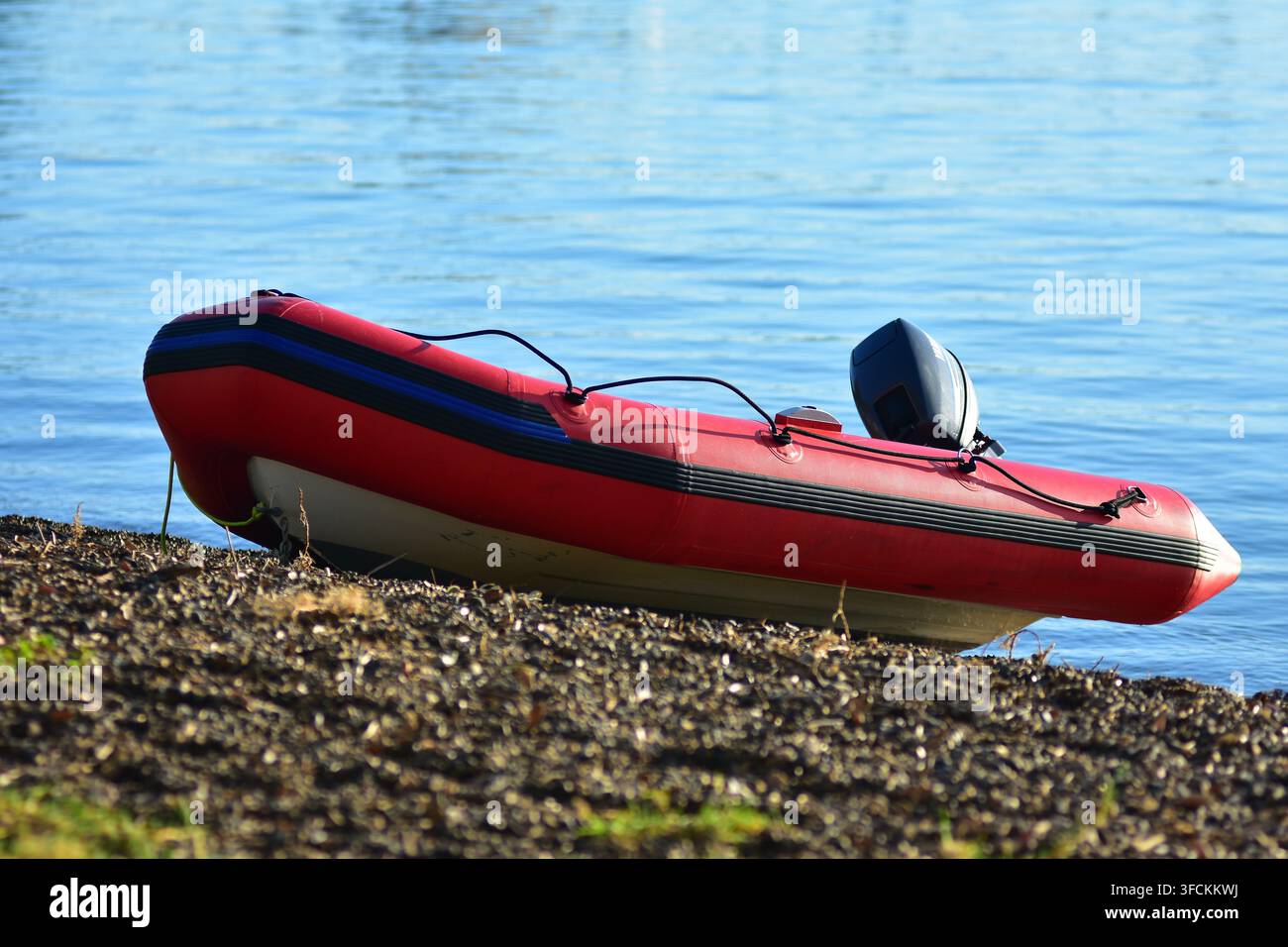 Ein rotes und weißes aufblasbares Sportdingi mit einem befestigten Außenbordmotor ruht an einem kiesigen Ufer am Wasserrand. Stockfoto