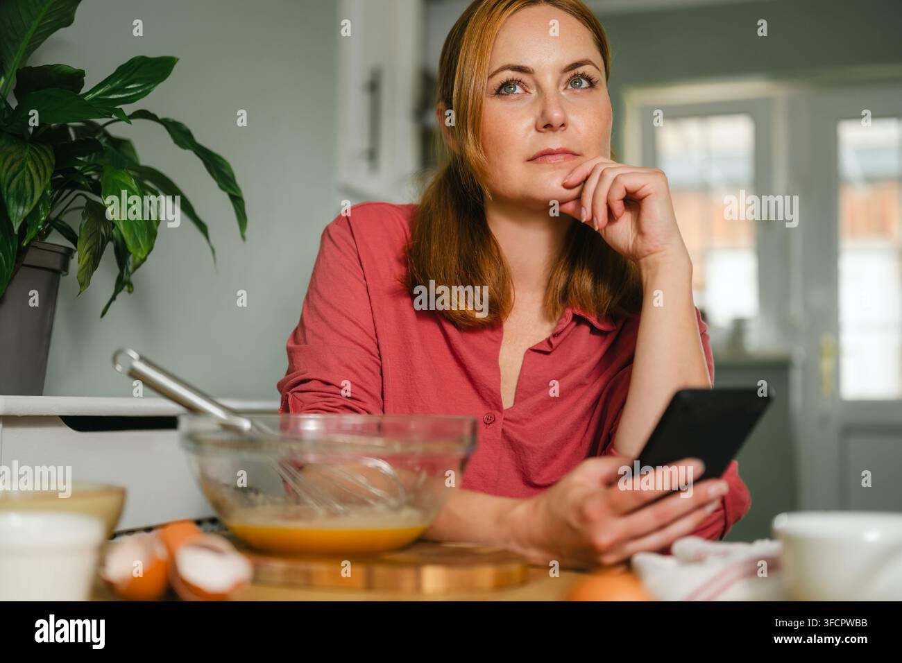 Nachdenkliche Frau schaut von Rezept auf dem Telefon auf, während sie backt Stockfoto