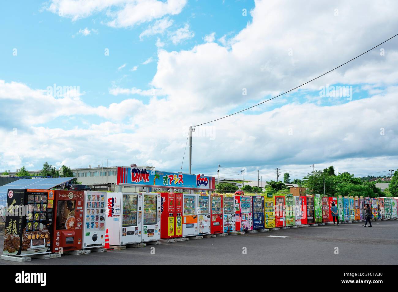 15. Juni 2025, Kurihara, Japan: Eine lange Reihe verschiedener Verkaufsautomaten, die im Freien auf einem Parkplatz unter einem bewölkten blauen Himmel stehen. Stockfoto