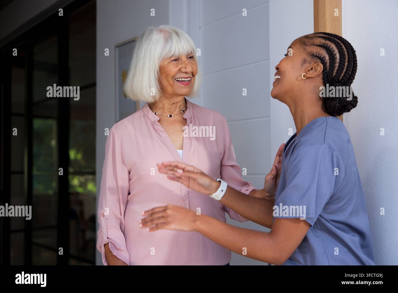 Afroamerikanische Pflegekraft und ältere Frau lächeln, begrüßen sich am Eingang mit Krankenschwesterkittel und Uhr Stockfoto