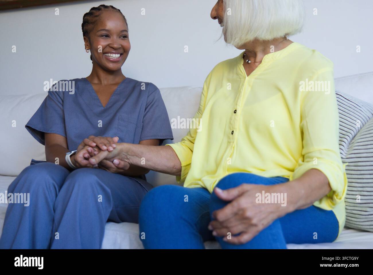 Afroamerikanische Pflegekraft und ältere Frau sitzen zu Hause auf der Couch und halten sich an den Händen Stockfoto