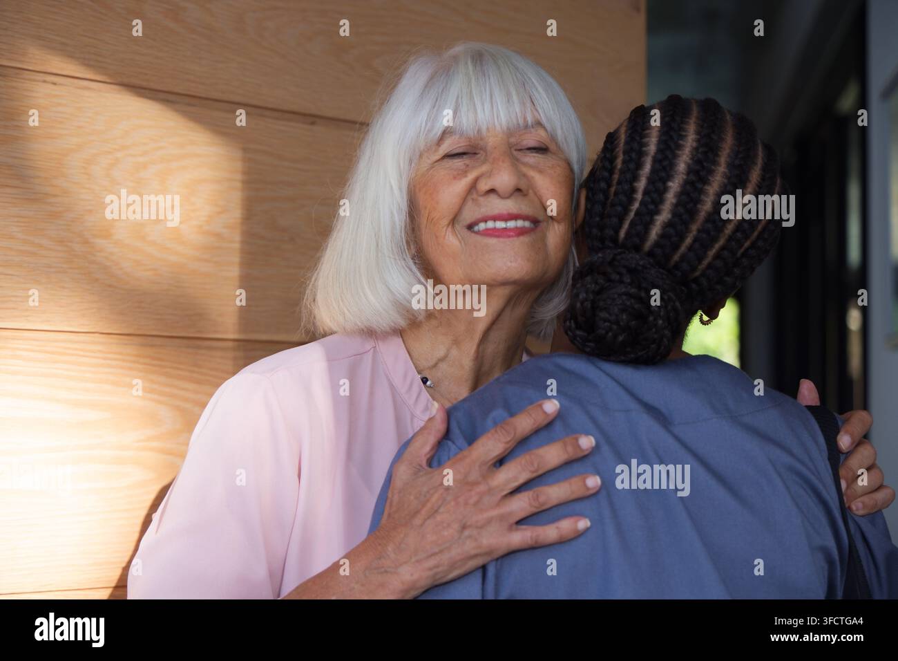 Seniorin und afroamerikanische Betreuerin umarmen sich in einem Pflegeflur, rosa Bluse und OP-Kleidung zu sehen Stockfoto