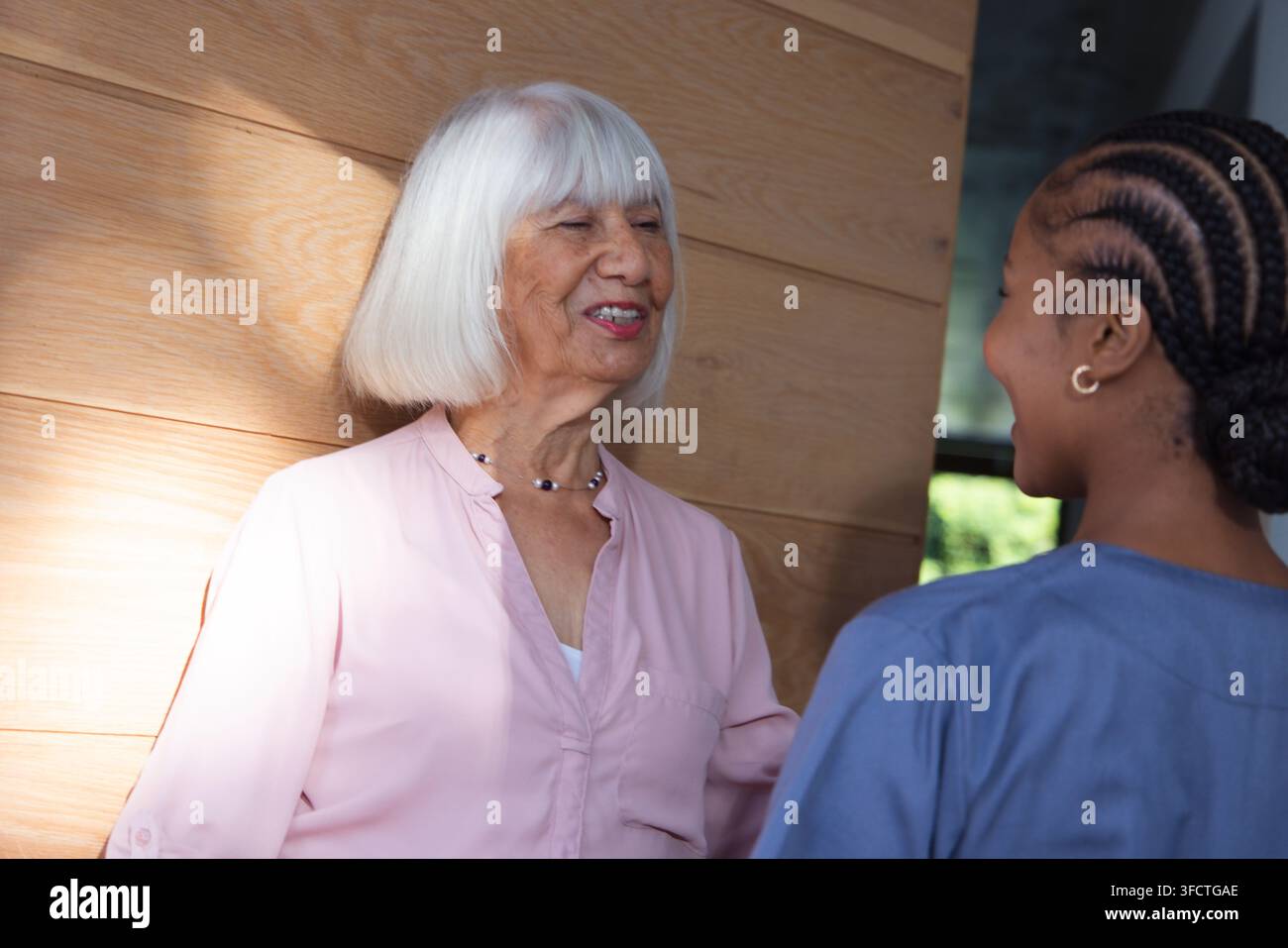 Seniorin und weibliche Betreuerin unterhalten sich in der Klinik an einer holzvertäfelten Wand bei Sonnenlicht Stockfoto