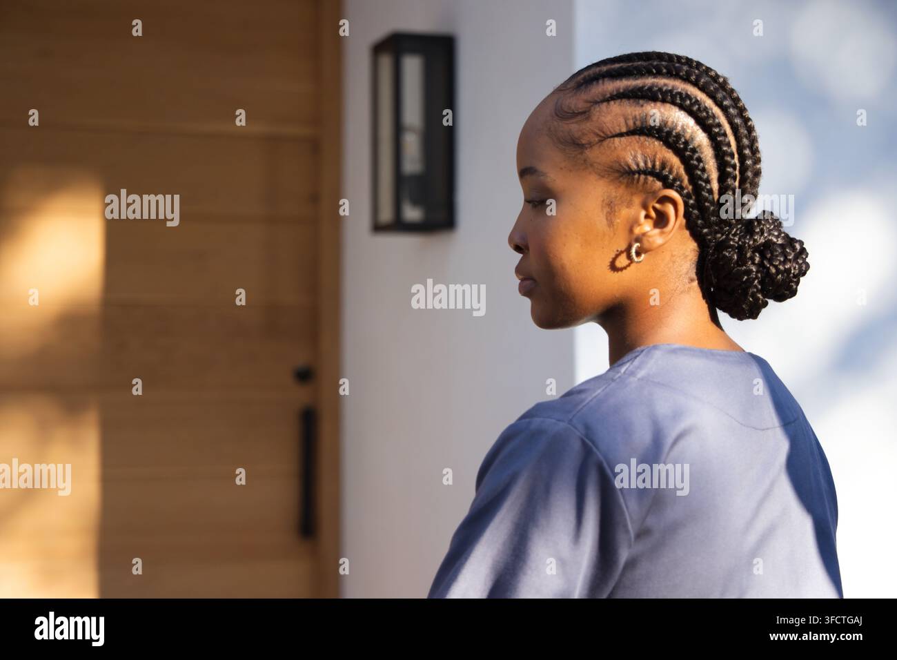 Erwachsene afroamerikanische Frau steht am Eingang und trägt blaue OP-Kleidung, Kreolen an der Holztür Stockfoto