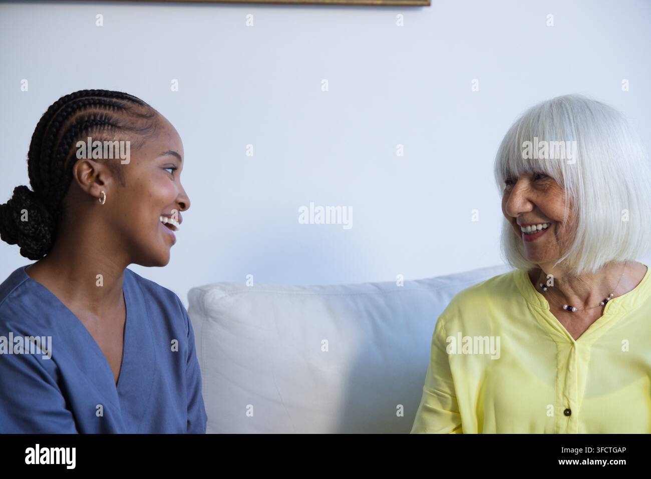 Afroamerikanische Pflegekraft im blauen Kittel spricht mit einer älteren Frau auf einem hellen Sofa im Wohnzimmer Stockfoto