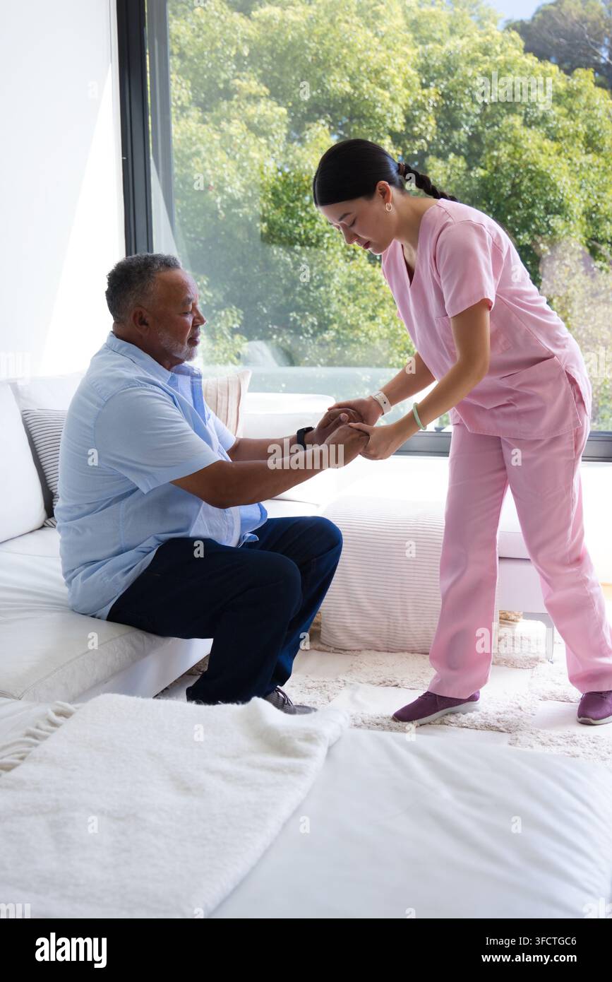 Asiatische Pflegekraft in rosa OP-Kleidung hilft einem afroamerikanischen Senioren, vom Sofa am Fenster aufzustehen Stockfoto