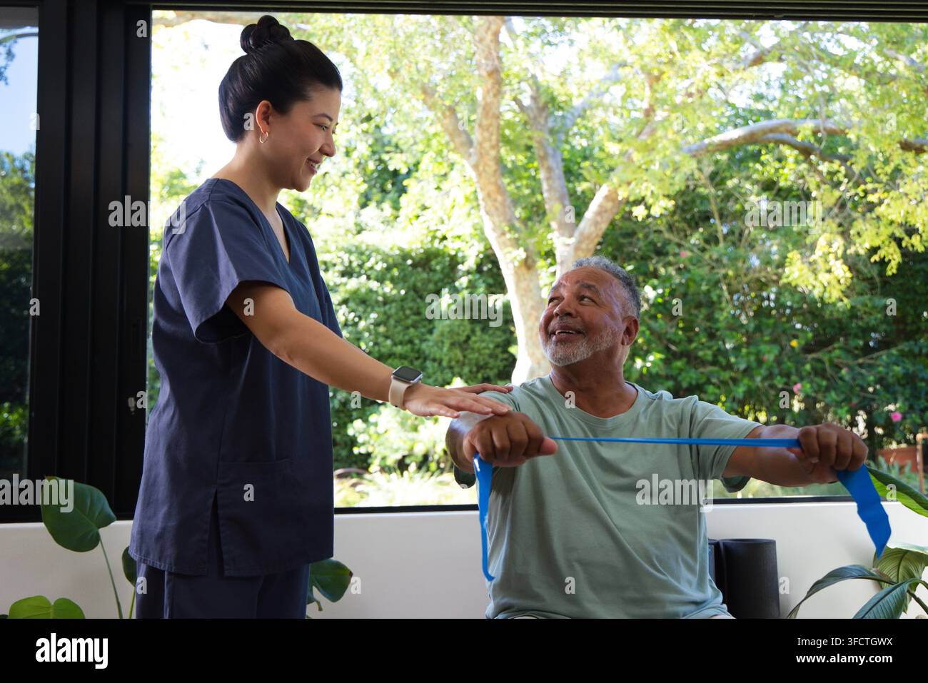 Asiatische Krankenschwester in marineblauen OP-Kitteln führt einen älteren afroamerikanischen Mann, der ein blaues Band in einem Therapieraum zieht Stockfoto