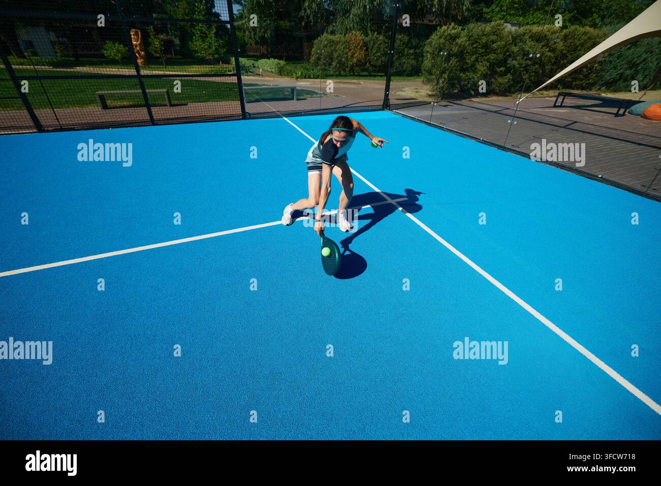 Maler Padel-Athlet in schwarzer Sportbekleidung, der sich dynamisch über einen minimalistischen blauen Außenplatz bewegt Stockfoto