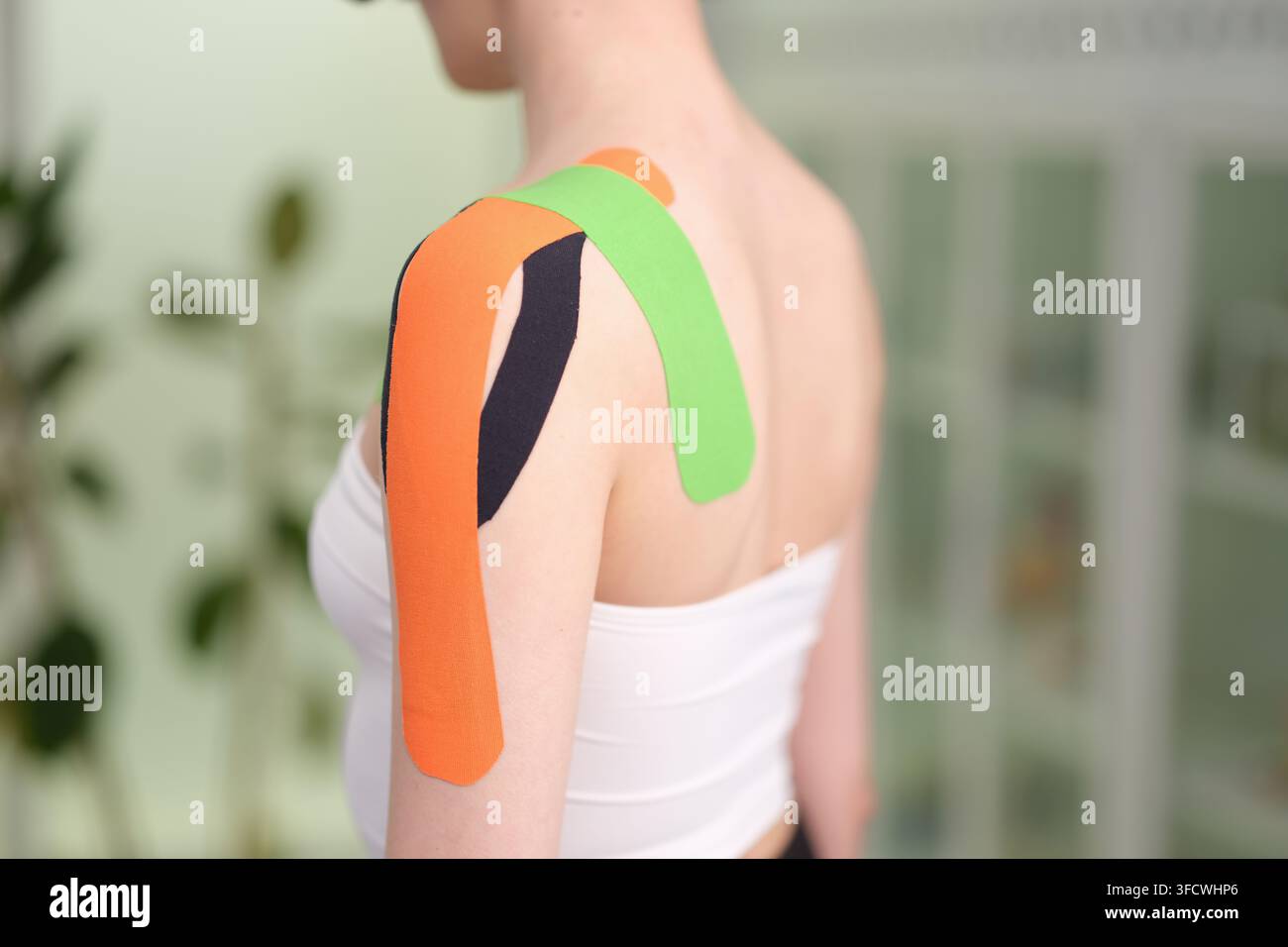 Frau, die Kinesiology-Tape zur Unterstützung oder Verletzungsbehandlung an der Schulter anbringt, in einer Indoor-Umgebung mit neutraler, klinischer Stimmung und Rückansicht der Frau Stockfoto