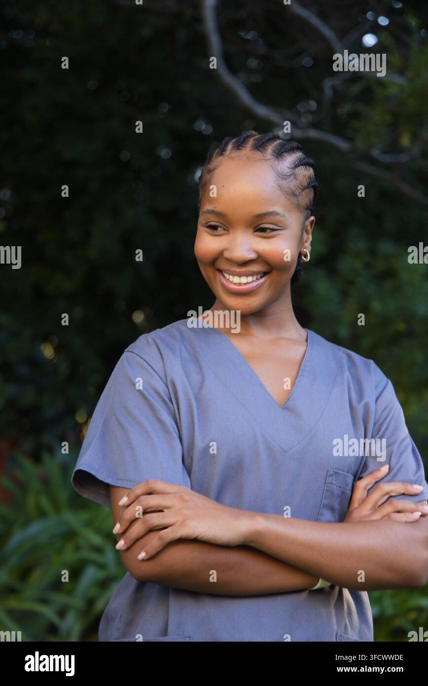 Frau lächelt und steht mit verschränkten Armen im Garten, trägt graublaue OP-Kleidung, Cornrows, Kreolen Stockfoto