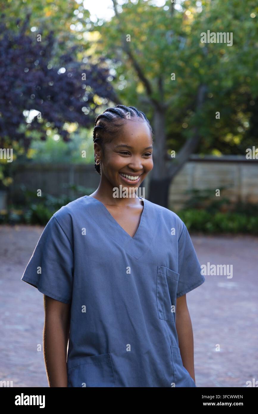 Afroamerikanische Frau steht lächelnd in einem gepflasterten Hinterhof und trägt blaue OP-Kleidung und geflochtene Cornrows Stockfoto