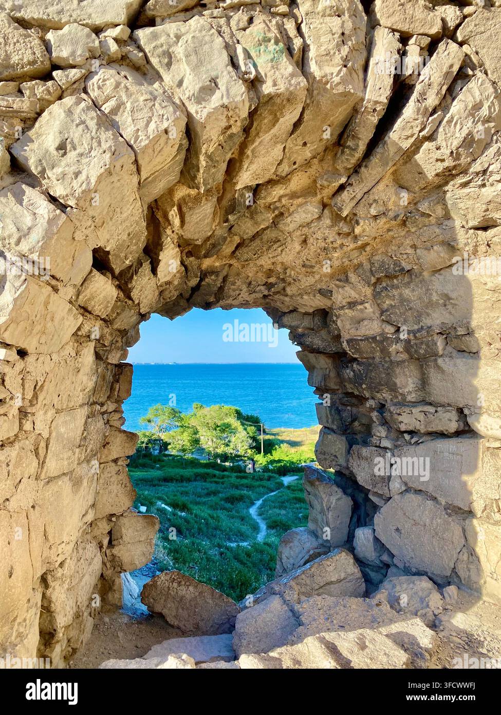Das blaue Meer erscheint durch eine alte Steinöffnung in einer sonnenbeschienenen Festungsmauer Stockfoto