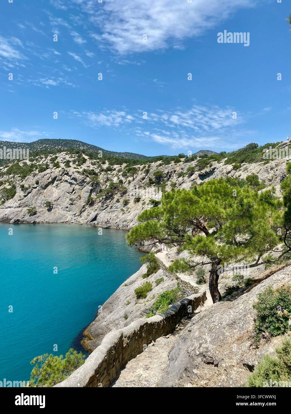 Steiniger Küstenpfad windet sich neben einer ruhigen türkisfarbenen Bucht im Sommersonnenschein - Smartphone-aufgenommenes Stockfoto