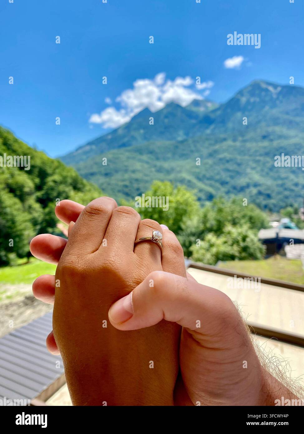 Paar hält sich an den Händen und zeigt einen Verlobungsring vor einer sonnigen Berglandschaft. - Smartphone-aufgenommenes Stockfoto