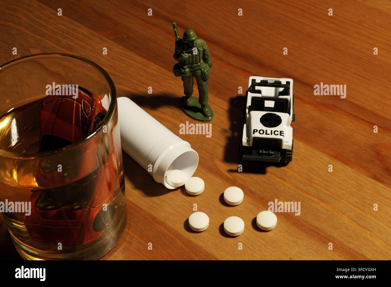 Kleines rotes Auto-Spielzeug, das in einem Glas Whiskey versinkt, neben verschütteten weißen Pillen aus einer Flasche, einem Spielzeugsoldaten und einem Spielzeug-Polizeiauto auf einem Holztisch, depi Stockfoto