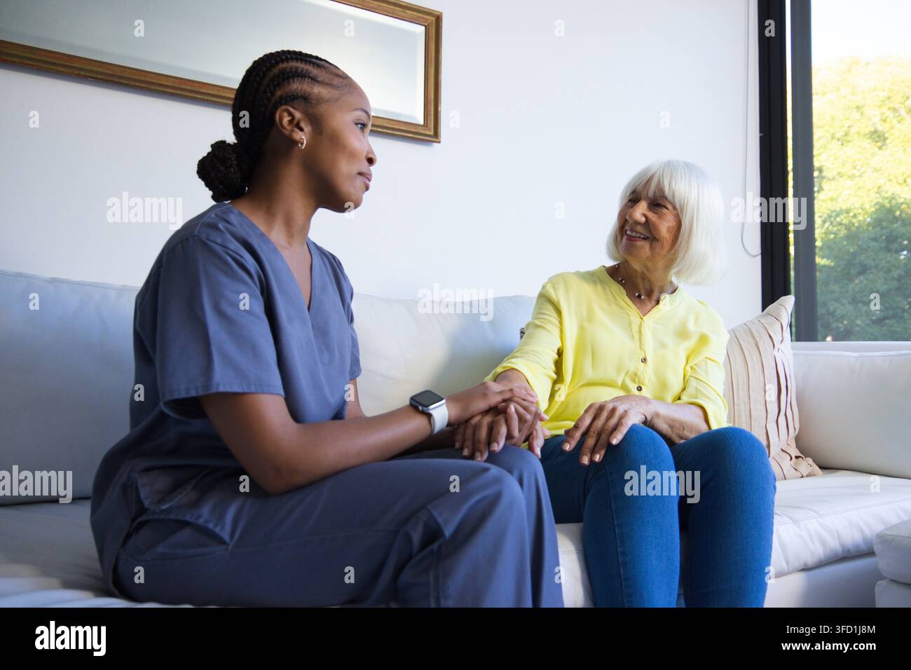 Vielfältige weibliche Pflegekraft in dunkelblauen Kitteln, die zu Hause auf dem Sofa sitzt und die Hand einer Seniorin hält Stockfoto