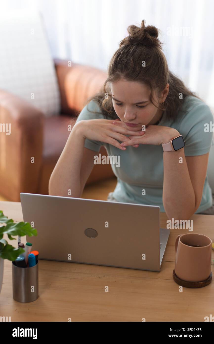 Frau lehnt sich nach vorne, konzentriert sich auf Laptop am Heimarbeitsplatz mit Tasse, Pflanze, Smartwatch Stockfoto