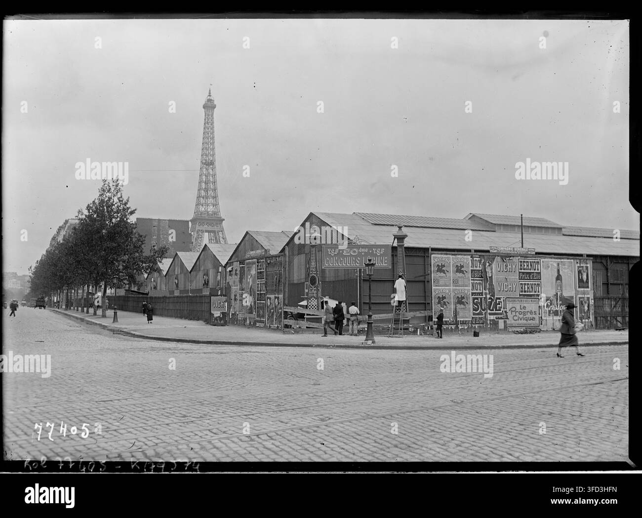 Eine Straßenszene in Paris mit dem Eiffelturm im Hintergrund, mit provisorischen Bauten und Werbetafeln entlang einer Kopfsteinpflasterstraße im Champ-de-Mars während des Lépine-Wettbewerbs von 1922 Stockfoto