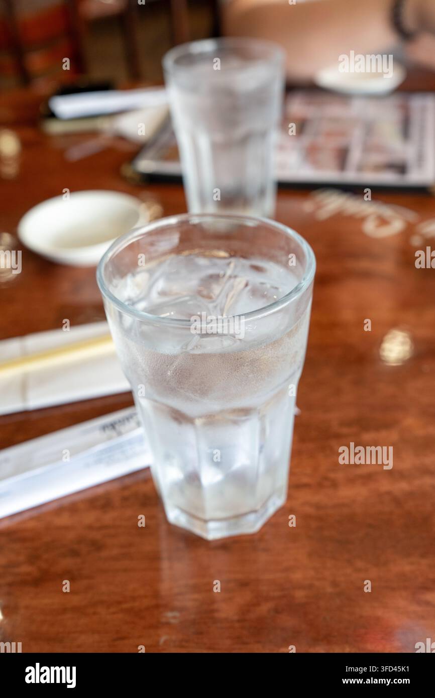 Nahaufnahme einer vertikalen Aufnahme eines klaren Glases gefüllt mit kaltem Wasser und Eiswürfeln auf einem hölzernen Restauranttisch. Kondensation am Glas und ein weiches Blau Stockfoto