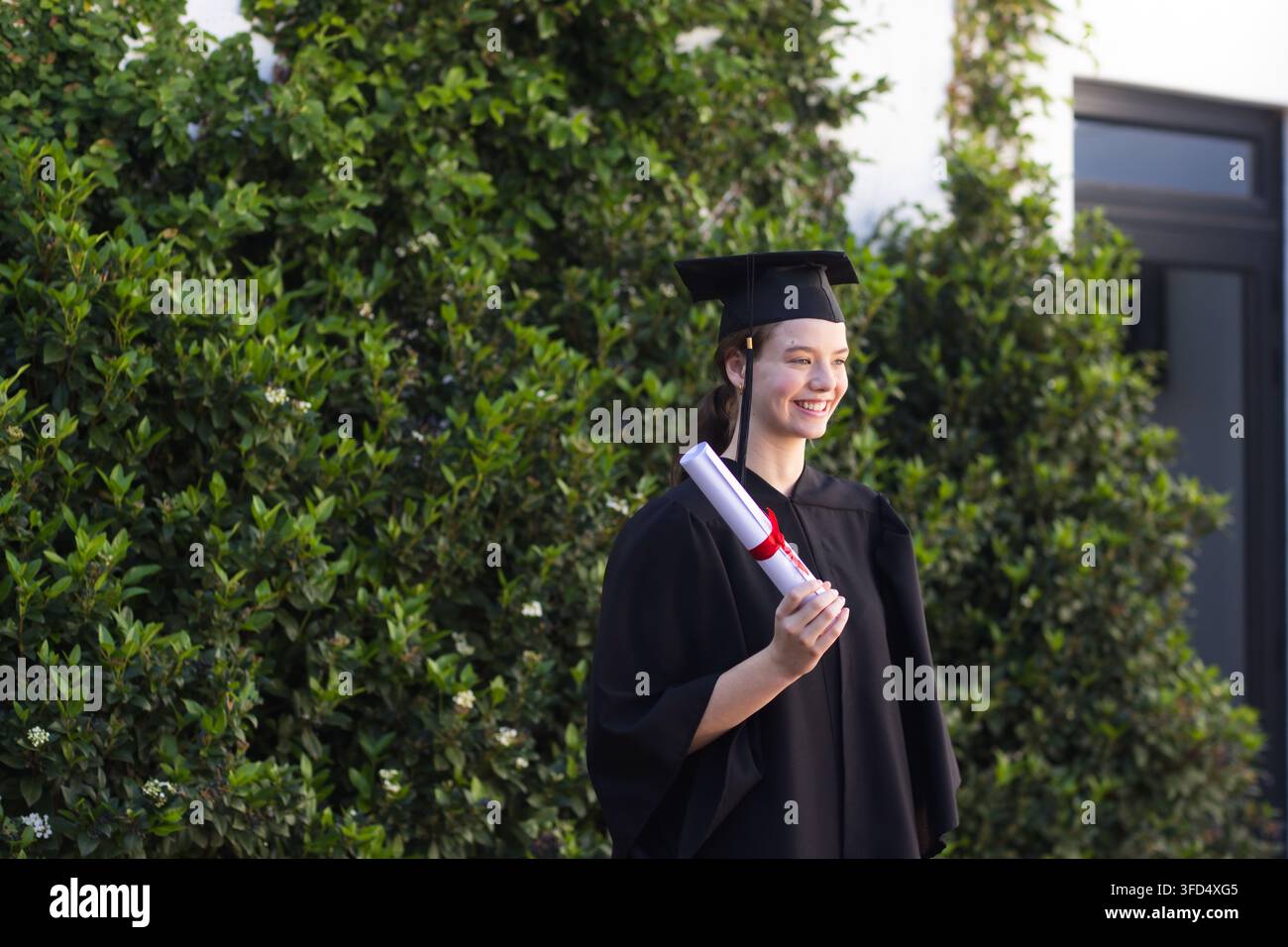 Absolventin steht auf dem Campus neben Sträuchern, trägt Robe und hält Diplom mit roter Schleife, Kopiraum Stockfoto