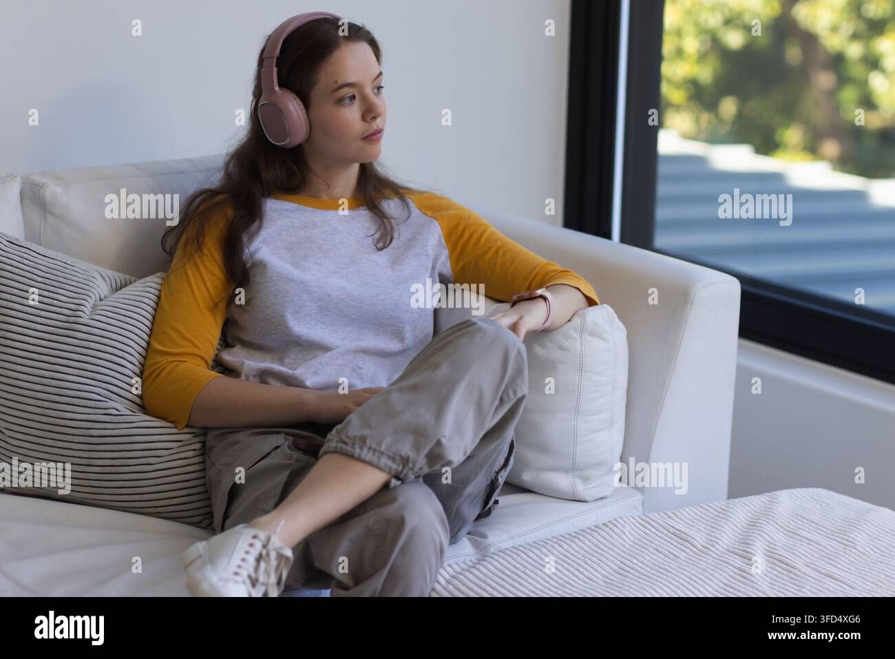 Frau Mitte 20, die auf einem hellen Ecksofa nahe einem großen Fenster sitzt, mit rosa Kopfhörern Stockfoto