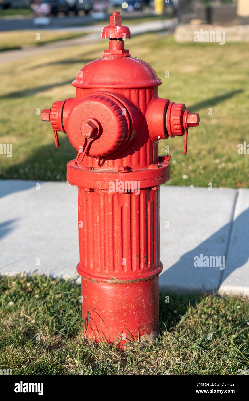 Nahaufnahme eines leuchtend roten Feuerhydranten, der an einem sonnigen Tag neben einem Gehweg und einem grasbewachsenen Wohngebiet steht. Stockfoto